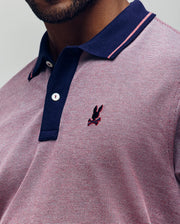 Close-up of a person wearing the Psycho Bunny QUINTON TEXTURE PIQUE POLO in pink and navy, featuring pink accents and an embroidered dark bunny logo on the chest. Only the lower face and upper torso are visible.