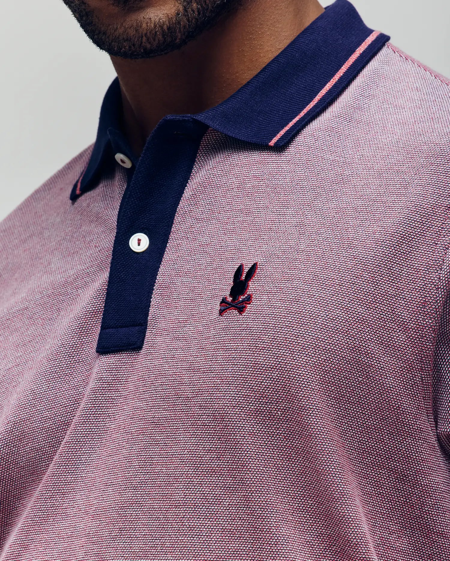 Close-up of a person wearing the Psycho Bunny QUINTON TEXTURE PIQUE POLO in pink and navy, featuring pink accents and an embroidered dark bunny logo on the chest. Only the lower face and upper torso are visible.