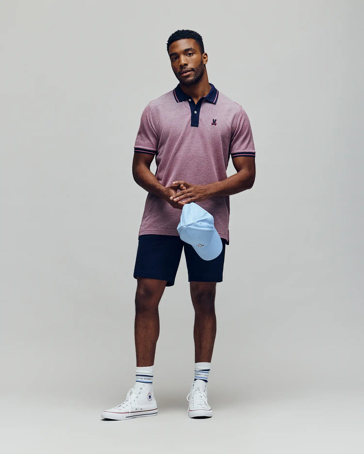 A man stands against a plain background, wearing the Psycho Bunny QUINTON TEXTURE PIQUE POLO in maroon, navy shorts, striped white socks, and white high-top sneakers. He holds a light blue baseball cap and looks calmly at the camera.