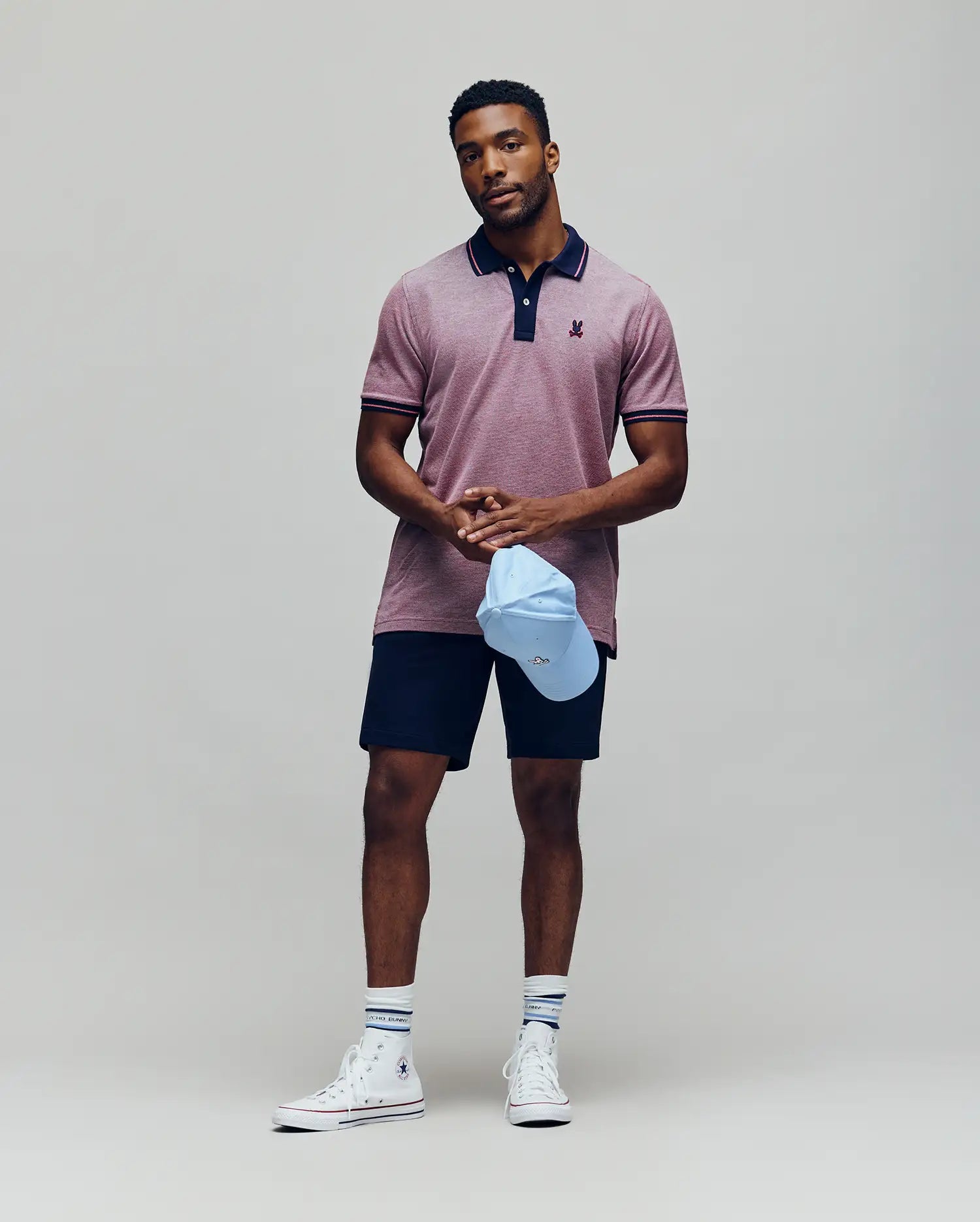 A man stands against a plain background, wearing the Psycho Bunny QUINTON TEXTURE PIQUE POLO in maroon, navy shorts, striped white socks, and white high-top sneakers. He holds a light blue baseball cap and looks calmly at the camera.