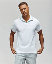 A man stands against a plain background, wearing the Psycho Bunny MENS JAXON SOFT PIQUE CAMP COLLAR POLO - MT0100006 in white with black trim and light gray pants, looking at the camera with a neutral expression, hands at his sides.