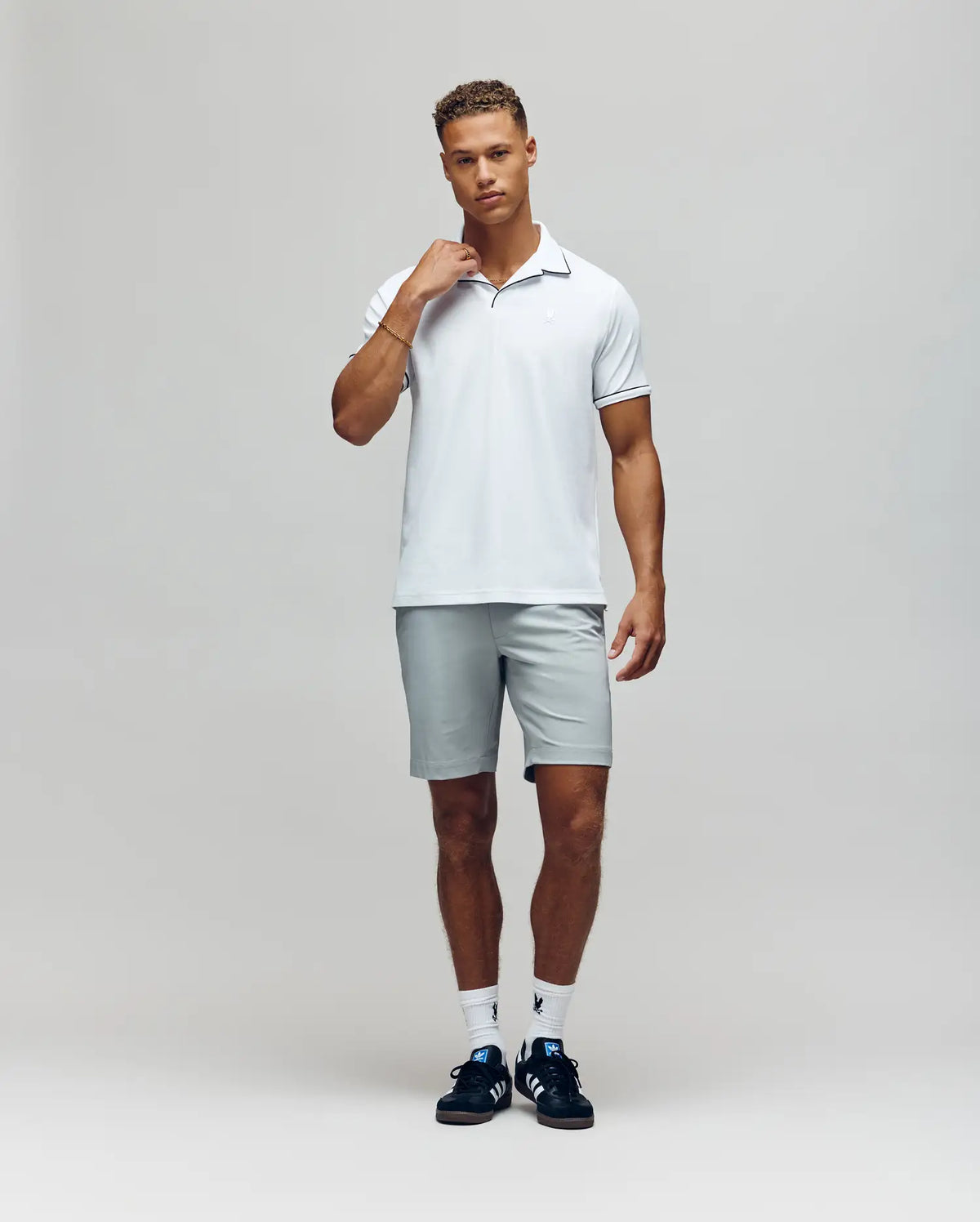 A man with short curly hair poses in a studio wearing the Psycho Bunny MENS JAXON SOFT PIQUE CAMP COLLAR POLO (MT0100006), light gray shorts, white socks, and black athletic shoes, with one hand near his neck as he looks at the camera.