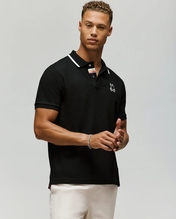 A man with short curly hair and a neutral expression stands against a light gray background, wearing light pants and the Psycho Bunny MENS FENLEY PIQUE POLO (MT0100009), a black Pima cotton polo with white trim and a small chest logo.