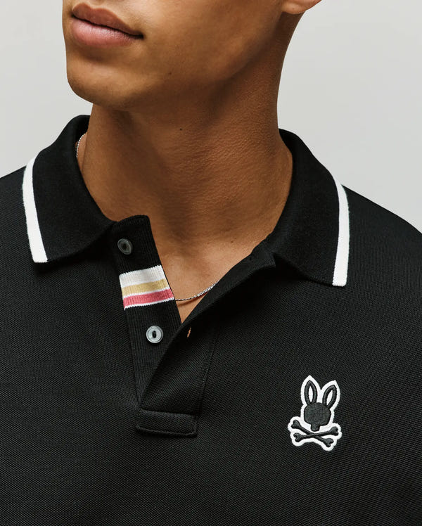 A person wears the Psycho Bunny Mens Fenley Pique Polo (MT0100009), a black Pima cotton shirt with white-trimmed collar, bunny skull-and-crossbones logo, and a placket featuring black buttons with beige, burgundy, and white stripes.