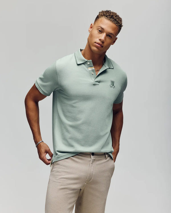 A man with short curly hair wears the Psycho Bunny Mens Fenley Pique Polo (MT0100009) in mint green, featuring an embroidered chest Bunny, paired with beige pants. He stands against a plain light background with one hand in his pocket.