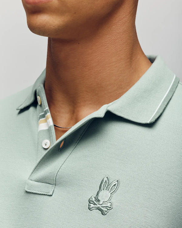 Close-up of a person wearing the Psycho Bunny MENS FENLEY PIQUE POLO (MT0100009) in light green Pima cotton, featuring white and yellow striped collar and embroidered Bunny logo. The face is partially out of frame.