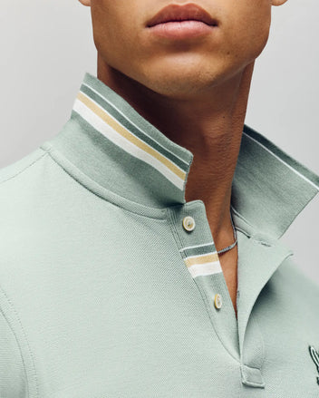 Close-up of a person wearing the Psycho Bunny MENS FENLEY PIQUE POLO (MT0100009) in light green Pima cotton with a beige and white striped collar, cream buttons, and an embroidered Bunny logo, styled with a thin chain necklace.
