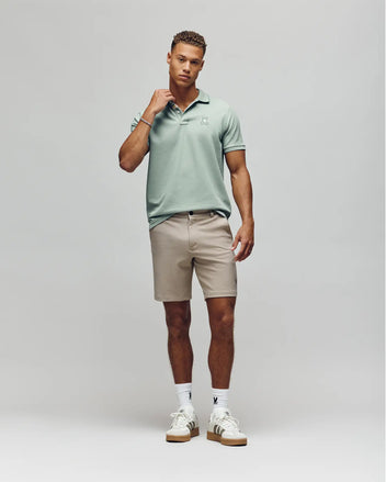 A man poses against a plain background in the Psycho Bunny MENS FENLEY PIQUE POLO - MT0100009 in light green, featuring an embroidered chest Bunny, paired with beige shorts and white sneakers.