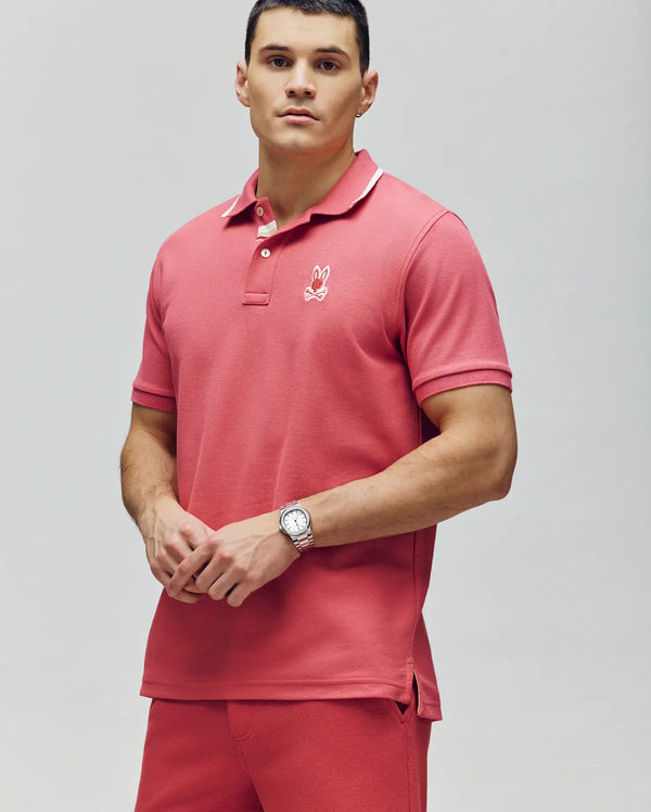 A man with short dark hair and a wristwatch wears a coral pink Psycho Bunny MENS FENLEY PIQUE POLO - MT0100009, made of soft Pima cotton with an embroidered chest Bunny logo, and matching shorts, standing against a light gray background.