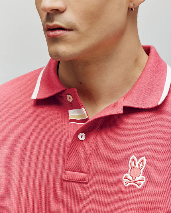 A person wears the Psycho Bunny MENS FENLEY PIQUE POLO (MT0100009) in pink, made from soft Pima cotton with an embroidered chest Bunny skull logo. The image shows their lower face, upper torso, and a small earring in the left ear.