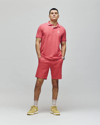 A man stands against a plain background wearing the Psycho Bunny MENS FENLEY PIQUE POLO (MT0100009) in pink Pima cotton with an embroidered chest Bunny, paired with matching pink shorts, yellow sneakers, and light-colored socks. Hands in pockets, he faces forward.