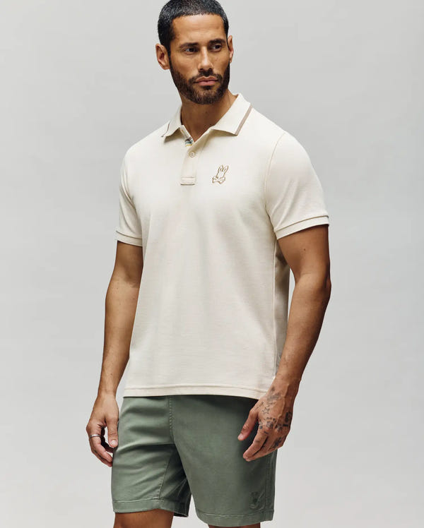 A man with short dark hair and a beard wears the Psycho Bunny MENS FENLEY PIQUE POLO - MT0100009 in beige, featuring Mother-of-Pearl buttons and an embroidered chest Bunny, paired with olive green shorts against a plain background.