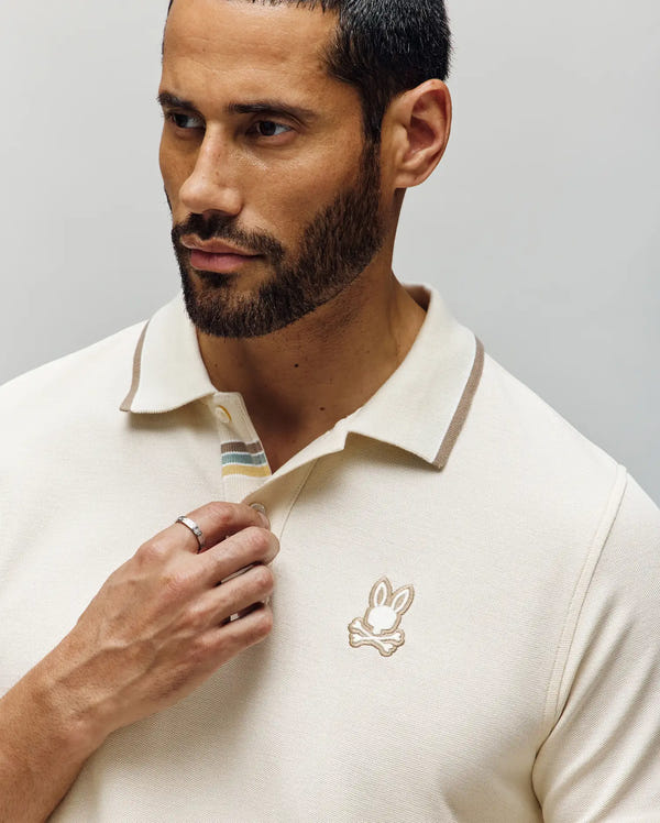 A man with short dark hair and a beard wears the Psycho Bunny MENS FENLEY PIQUE POLO (MT0100009) in cream, featuring an embroidered Bunny logo on the chest. He looks to the side while adjusting the collar with his left hand.