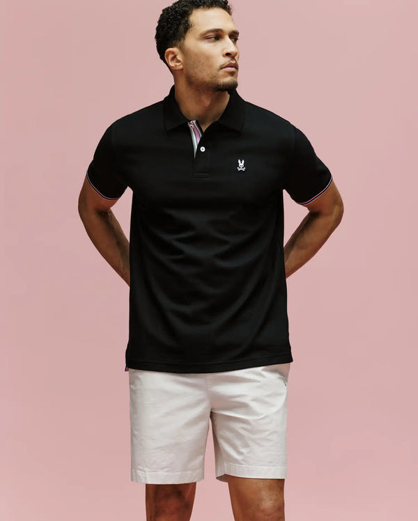 A man stands against a plain pink background wearing the Psycho Bunny MENS ARCHIVE CLASSIC BUNNY PIQUE POLO (MT0100010) in black, featuring an embroidered Bunny logo on the chest, paired with light shorts. His hands are behind his back as he looks to the side.