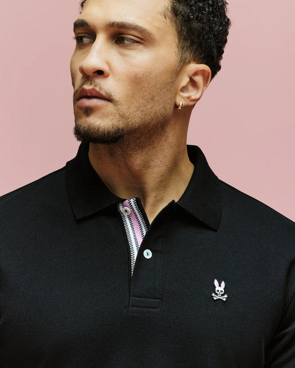 A man with short curly hair and a trimmed beard wears the Psycho Bunny MENS ARCHIVE CLASSIC BUNNY PIQUE POLO (MT0100010) in black, featuring an embroidered Bunny logo on the chest, standing against a pink background and looking left.