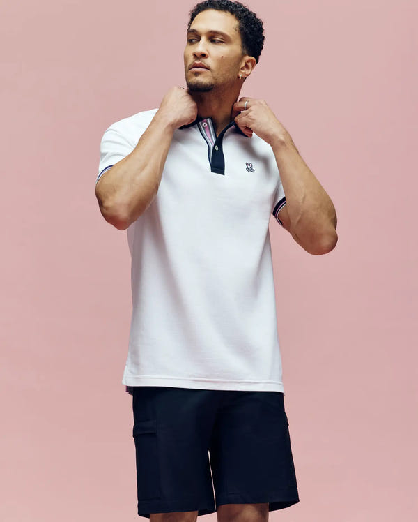 A man stands against a pink background, adjusting the collar of his Psycho Bunny MENS ARCHIVE CLASSIC BUNNY PIQUE POLO - MT0100010 with black and red trim, paired with black shorts.
