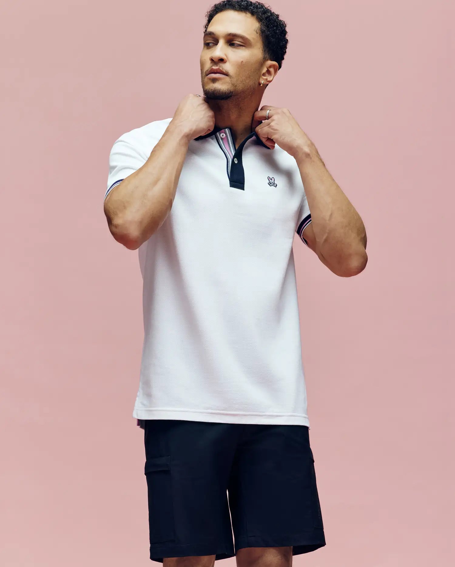 A man stands against a pink background, adjusting the collar of his Psycho Bunny MENS ARCHIVE CLASSIC BUNNY PIQUE POLO - MT0100010 with black and red trim, paired with black shorts.