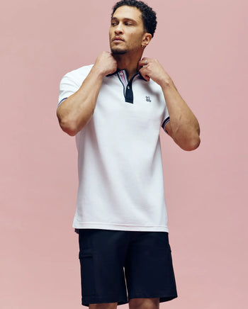 A man stands against a pink background, adjusting the collar of his Psycho Bunny MENS ARCHIVE CLASSIC BUNNY PIQUE POLO - MT0100010 with black and red trim, paired with black shorts.