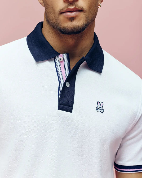 A person wears the Psycho Bunny MENS ARCHIVE CLASSIC BUNNY PIQUE POLO (MT0100010)—a white Pima cotton polo with a navy collar and embroidered Bunny logo—against a pink background, cropped at the nose and below the arms.