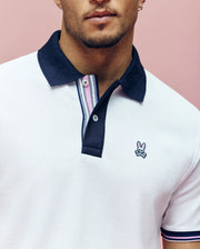 A person wears the Psycho Bunny MENS ARCHIVE CLASSIC BUNNY PIQUE POLO (MT0100010)—a white Pima cotton polo with a navy collar and embroidered Bunny logo—against a pink background, cropped at the nose and below the arms.