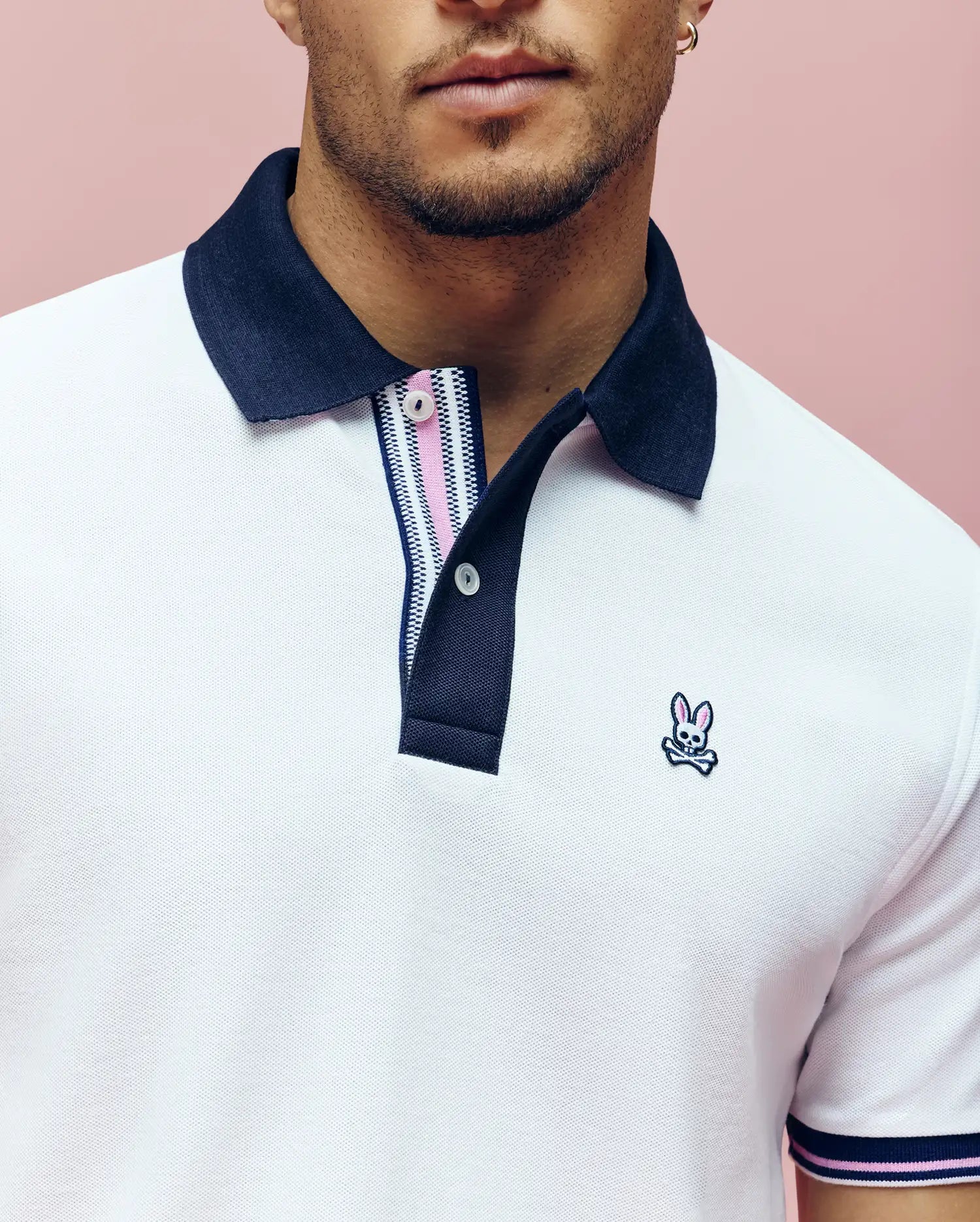 A person wears the Psycho Bunny MENS ARCHIVE CLASSIC BUNNY PIQUE POLO (MT0100010)—a white Pima cotton polo with a navy collar and embroidered Bunny logo—against a pink background, cropped at the nose and below the arms.