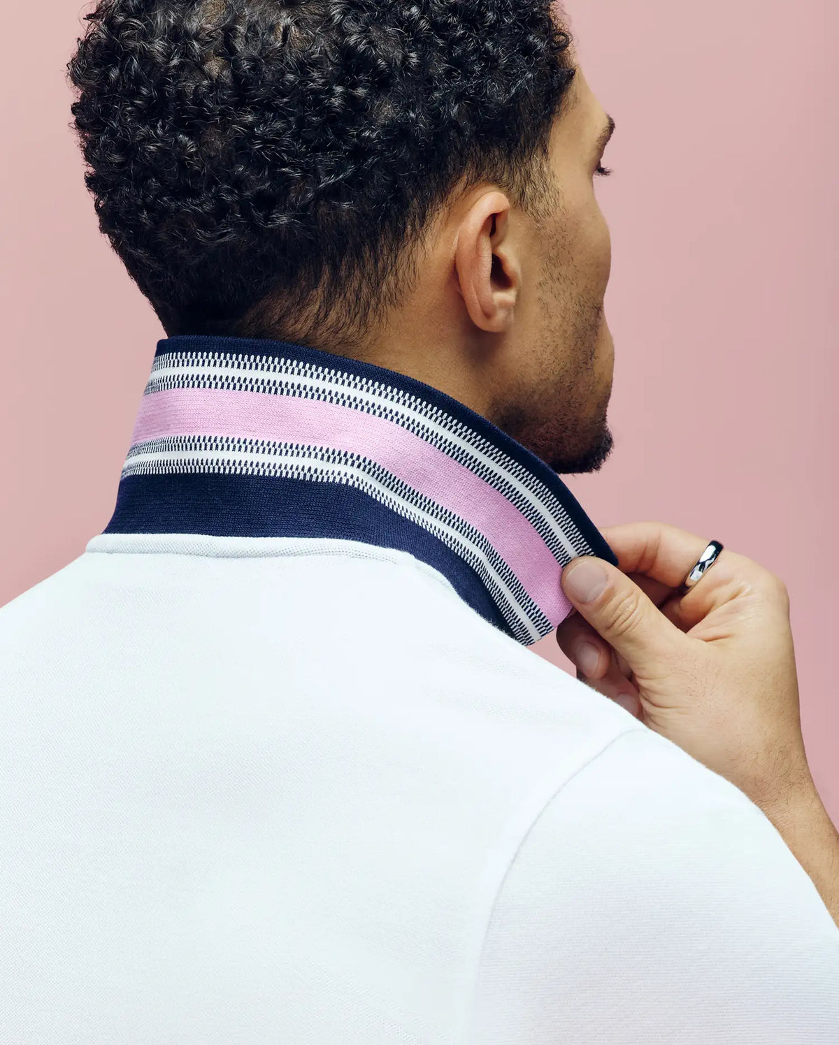 A man with short curly hair, seen from behind, lifts the collar of his white Psycho Bunny MENS ARCHIVE CLASSIC BUNNY PIQUE POLO (MT0100010) to reveal a navy, pink, and white striped underside against a plain pink background.