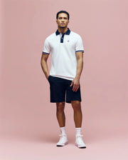 A man stands against a pink background wearing the Psycho Bunny MENS ARCHIVE CLASSIC BUNNY PIQUE POLO - MT0100010 with a dark collar, navy shorts, white socks, and white high-top sneakers.