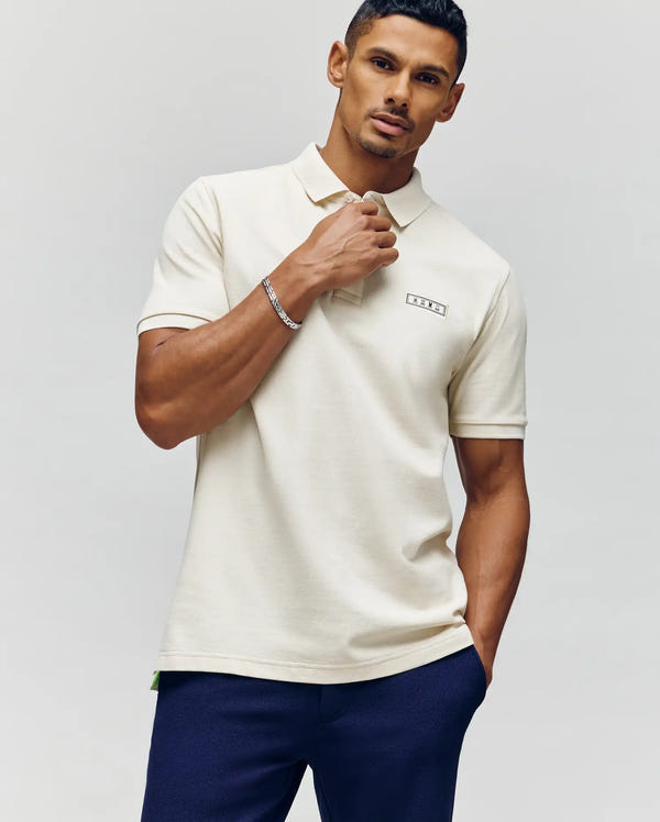 A man stands against a neutral background wearing the Psycho Bunny MENS AXTON PIQUE POLO - MT0100011 in cream with dark blue pants. He has short, styled hair and poses confidently, left hand near his collar, looking at the camera.