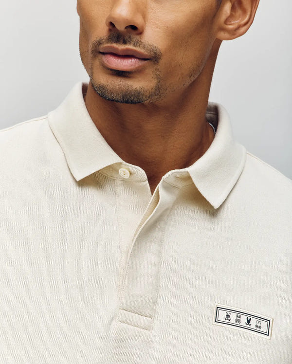 A man wears the Psycho Bunny MENS AXTON PIQUE POLO (MT0100011) in cream, made from Pima cotton with a collar and a small jacquard-woven logo on the chest. Only his lower face and upper torso are visible against a plain background.