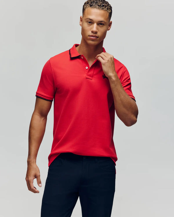 A young man with short, curly hair wears the Psycho Bunny MENS JUDE PIQUE POLO - MT0100012 in bright red and dark pants, standing against a plain light gray background and looking directly at the camera with a neutral expression.