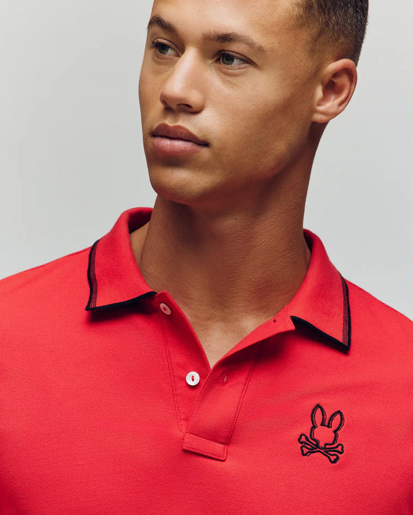 A young man models the Psycho Bunny MENS JUDE PIQUE POLO (MT0100012), a bright red Pima cotton shirt with black collar trim and the signature black bunny skull logo, posing against a plain light background.