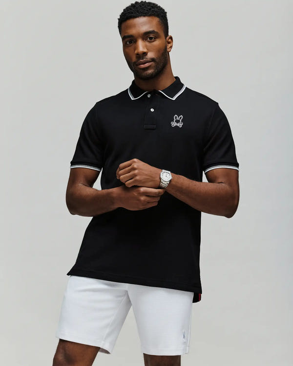 A man in the Psycho Bunny MENS JUDE PIQUE POLO - MT0100012, a black Pima cotton polo with white trim and bunny logo, pairs it with white shorts. He stands against a plain light background, wearing a wristwatch and has short hair.