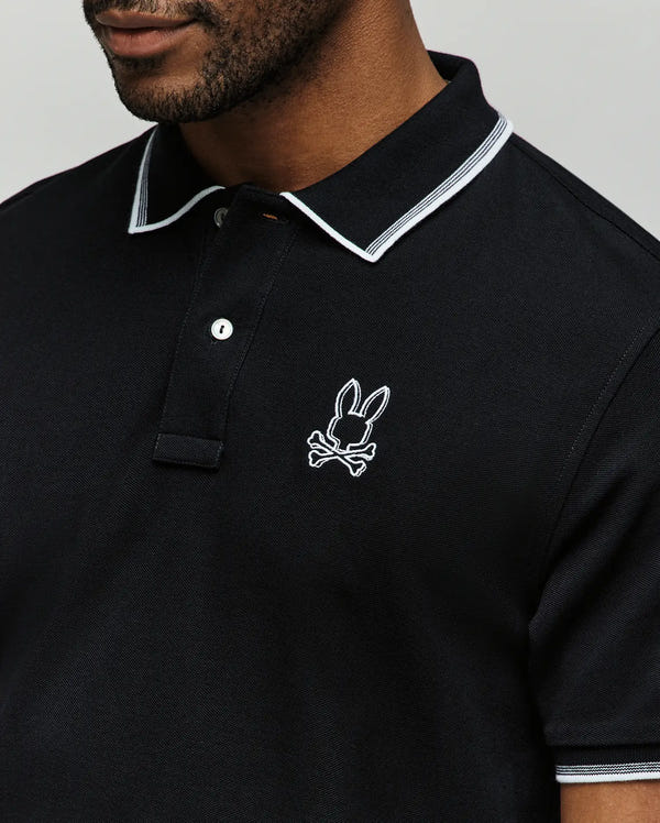 A man wears the Psycho Bunny MENS JUDE PIQUE POLO (MT0100012) in black with white collar trim, made from soft Pima cotton and featuring an embroidered Bunny head above crossed bones on the chest.