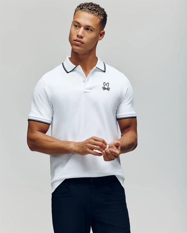 A man stands against a plain background wearing the Psycho Bunny MENS JUDE PIQUE POLO - MT0100012, featuring soft Pima cotton, black trim, and an embroidered bunny logo on the chest. He pairs it with dark pants and looks to the side with hands loosely clasped.