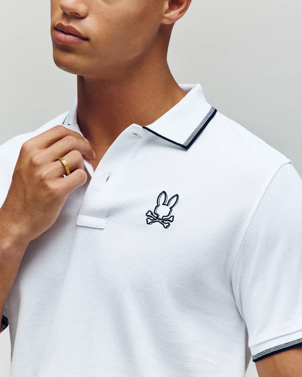 Someone wearing the Psycho Bunny MENS JUDE PIQUE POLO - MT0100012 in white, made from soft Pima cotton with a black embroidered bunny and crossbones on the chest, black trim, touches the collar while sporting a gold ring.