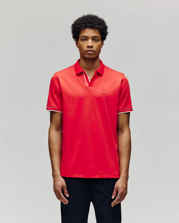 A man with short curly hair wears the Psycho Bunny MENS KENDELL INTERLOCK JOHNNY COLLAR POLO (MT0100015) in bright red with white-trimmed collar and sleeves, standing against a plain light gray background.