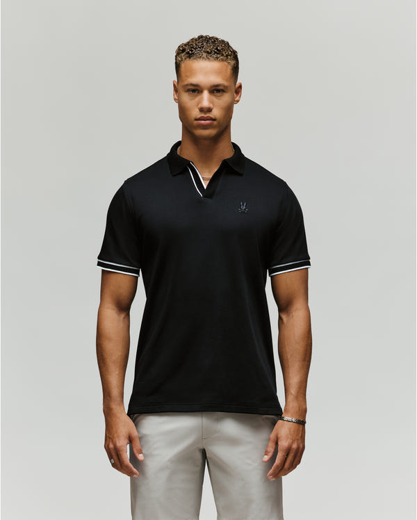 A man faces forward against a plain background wearing the Psycho Bunny MENS KENDELL INTERLOCK JOHNNY COLLAR POLO (MT0100015) in black with white trim, paired with light-colored pants. His arms rest at his sides and he has a neutral expression.
