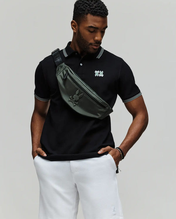 A man wears the Psycho Bunny MENS PB ARCHIVE NEON BUNNY POLO (MT0100018) in black Pima cotton with white trim, paired with white shorts and a dark green crossbody bag, standing hands-in-pockets against a light background. Made in Peru.