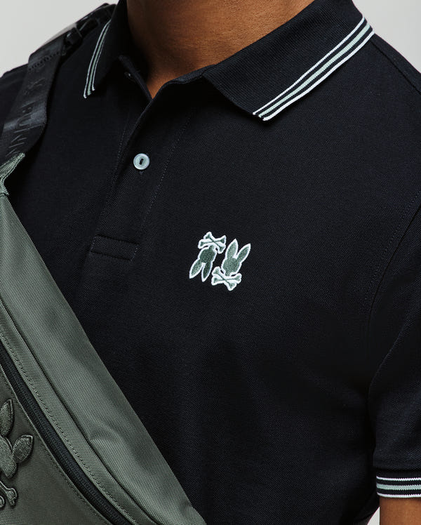 A person wears the Psycho Bunny MENS PB ARCHIVE NEON BUNNY POLO (MT0100018), a black 100% Pima cotton shirt with white-striped collar and embroidered Bunny logo, carrying a gray crossbody bag with matching logo. Made in Peru.