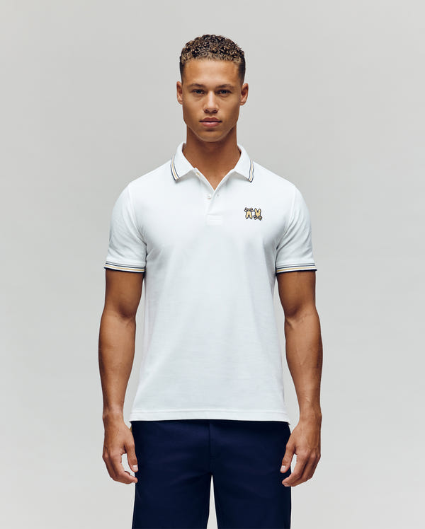 A young man poses against a plain light background, wearing Psycho Bunny’s MENS ARCHIVE NEON BUNNY POLO (MT0100018), featuring white Pima cotton, striped collar and sleeves, navy pants, and an embroidered Bunny logo on the chest.