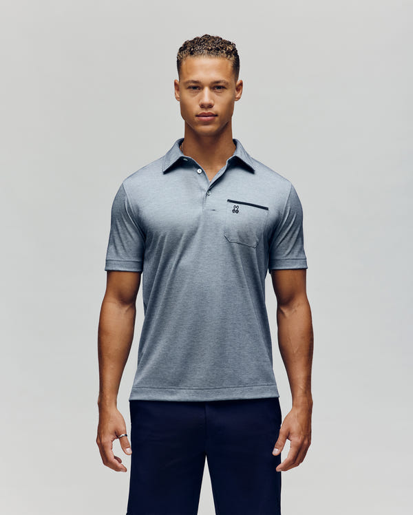 A man faces forward in a Psycho Bunny MENS HADLEY PERFORMANCE PIQUE POLO - MT0100021, light gray with a chest pocket, paired with dark navy pants, standing against a plain light background.