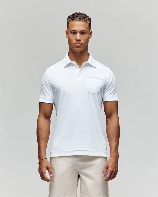 A man with short curly hair and a neutral expression stands facing forward against a plain light gray background, wearing the Psycho Bunny MENS HADLEY PERFORMANCE PIQUE POLO (MT0100021) in white and light-colored shorts.