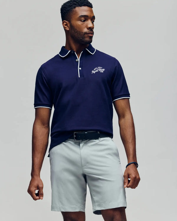 A man poses against a plain background wearing the Psycho Bunny MENS MATTEO PIQUE POLO (MT0100024) in navy with white trim and embroidered logo, paired with light gray shorts and a black belt. His head is slightly turned left, arms relaxed.