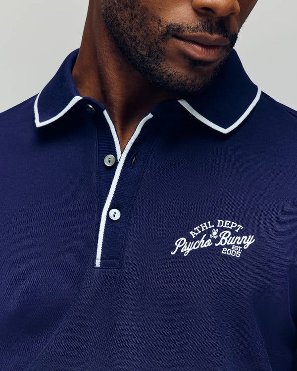 A man wears the Psycho Bunny MENS MATTEO PIQUE POLO - MT0100024 in navy blue, featuring white trim, Mother-of-Pearl buttons, and an "ATHL DEPT Psycho Bunny Est. 2005" embroidered logo on the left chest; only his lower face and torso are visible.