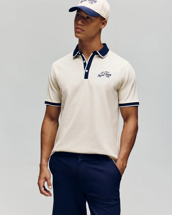 A man wears the Psycho Bunny MENS MATTEO PIQUE POLO - MT0100024 in cream with navy trim and Mother-of-Pearl buttons, navy pants, and a matching cream and navy cap. He stands with one hand in his pocket against a plain light gray background.