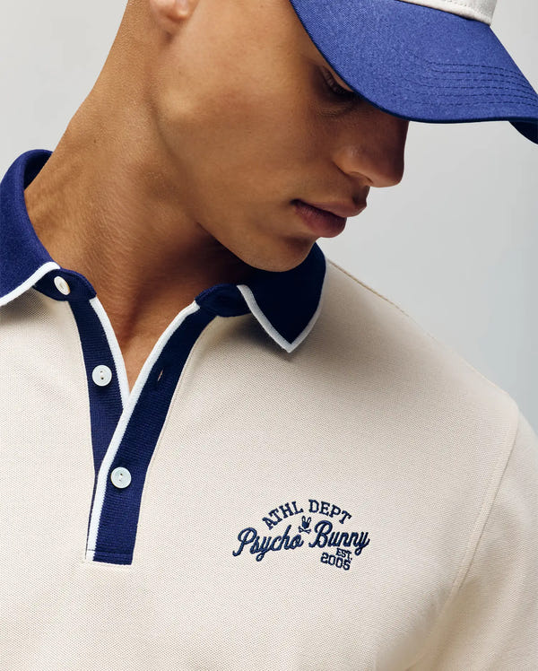 A man in a Psycho Bunny MENS MATTEO PIQUE POLO - MT0100024, cream Pima cotton with blue collar, trim, "ATHL DEPT Psycho Bunny EST 2005" logo embroidery, mother-of-pearl buttons, and a matching blue-white cap looks downward.