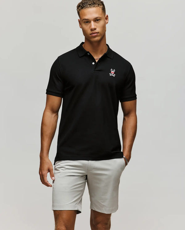 A man wears a black Psycho Bunny MENS MISCHIEF BUNNY PIQUE POLO (MT0100025) featuring mother-of-pearl buttons and a small chest logo, paired with light gray shorts, standing with one hand in his pocket against a plain light background.