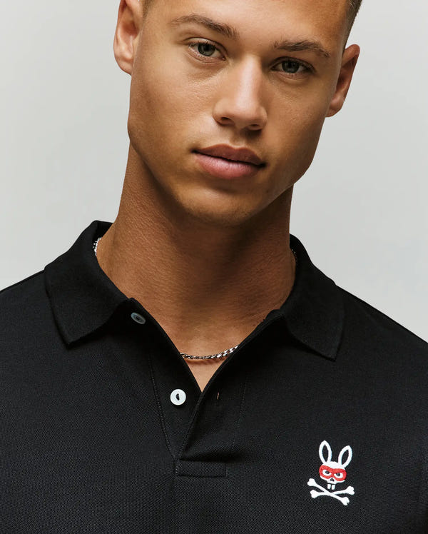 A young man in a Psycho Bunny MENS MISCHIEF BUNNY PIQUE POLO (MT0100025), featuring Pima cotton and a small bunny and crossbones logo on the chest, looks at the camera against a plain light background.