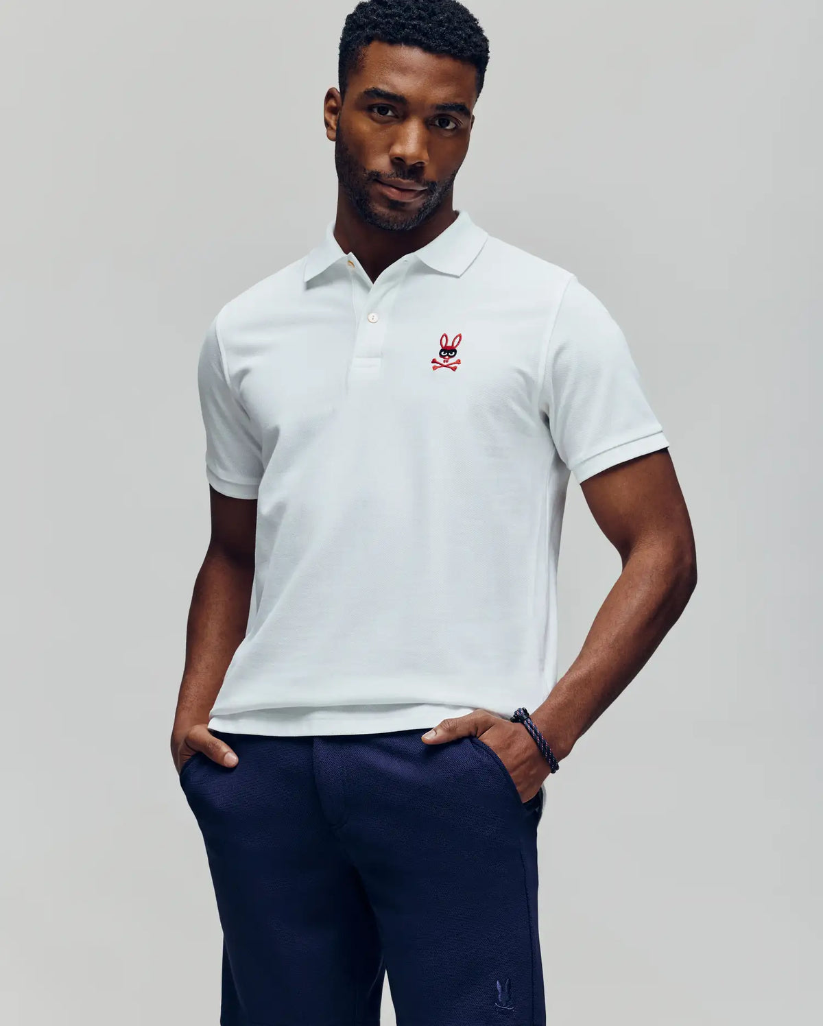A man wears a white Psycho Bunny Mens Mischief Bunny Pique Polo (MT0100025) with mother-of-pearl buttons and a red embroidered logo, paired with navy pants, hands in pockets against a plain background.
