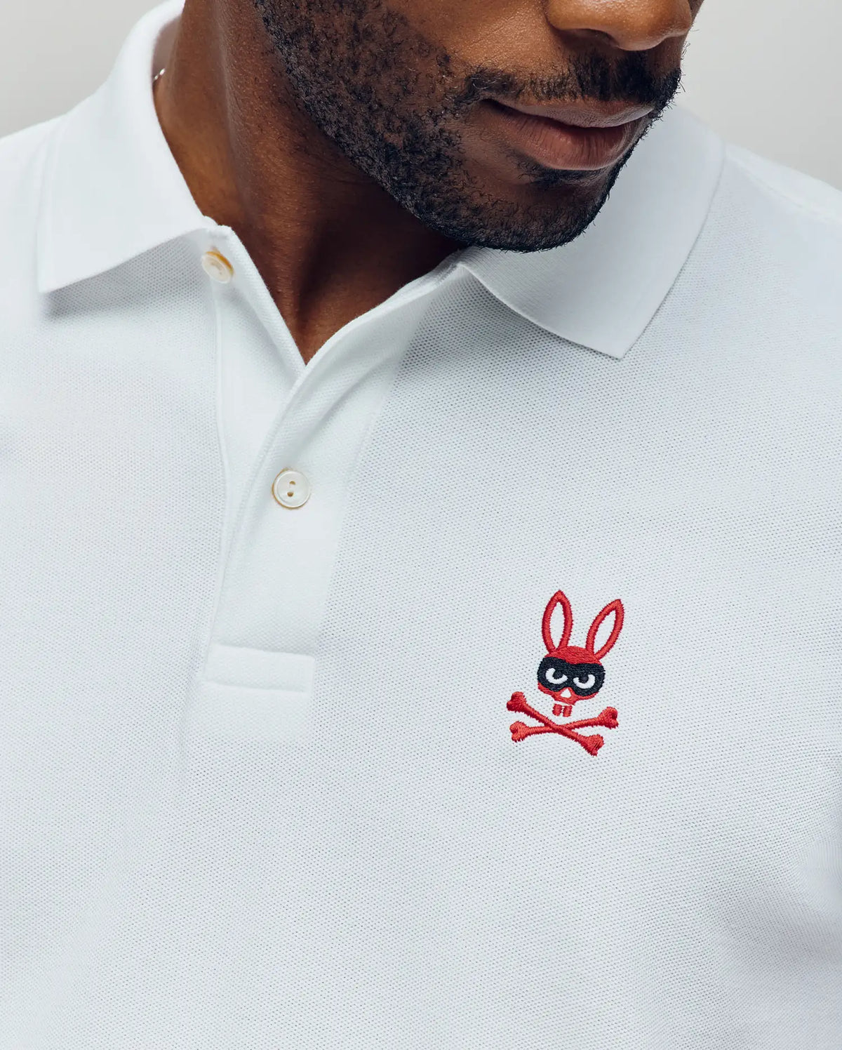 A man wears the Psycho Bunny MENS MISCHIEF BUNNY PIQUE POLO (MT0100025), made from soft Pima cotton with a red and black cartoon bunny and crossbones logo. Only his upper torso and lower face are visible.